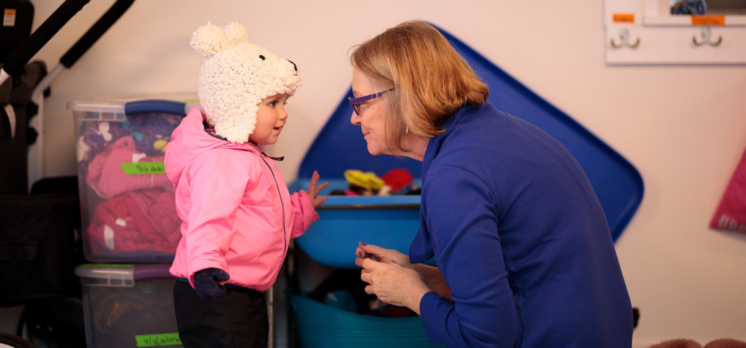 A toddler in winter coat and hat talks to a smiling early childhood educator