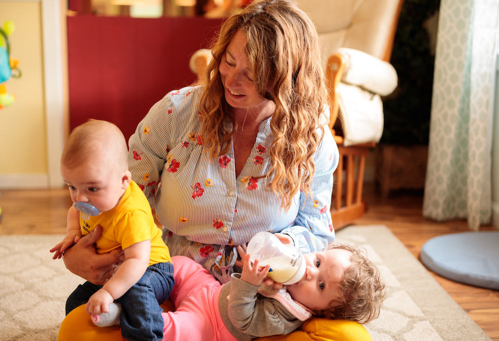 In a sunlit room, a woman with light skin, long curly red hair, and a blue flowered shirt smiles crosslegged holding two babies on her lap. A curly haired baby lies down and drinks from a bottle looking at the viewer, a baby in a yellow shirt and blue pacifier sits up ready to wiggle.