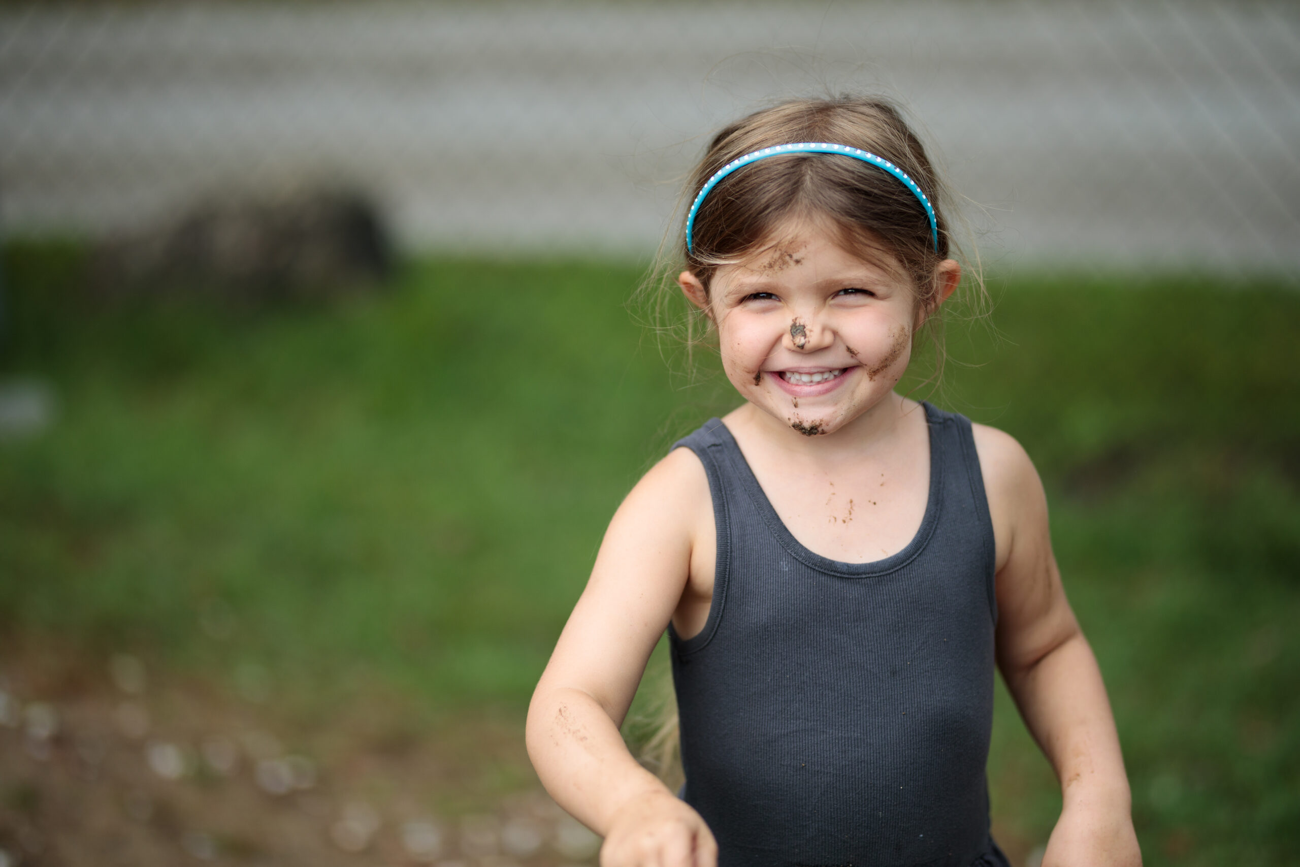 Little girl with gray tank top, blue headband, light skin, long brown hair stands outside and smiles with a big mud smudge on her nose