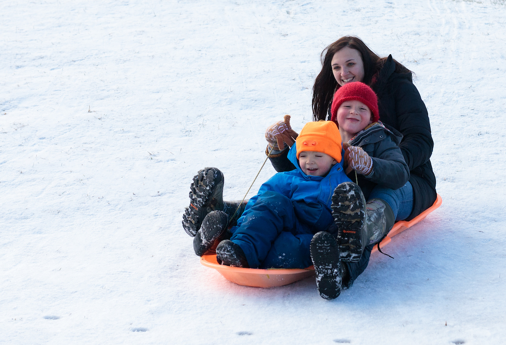 Three people ride down a snowy hill on an orange plastic sled. Two smiling children in orange and red hats are in the front and middle, a smiling teacher with long brown hair is in the back.