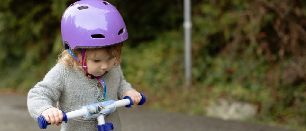 A small child with a determined expression wearing a light purple helmet and gray onesie pushes a scooter down a wooded road.
