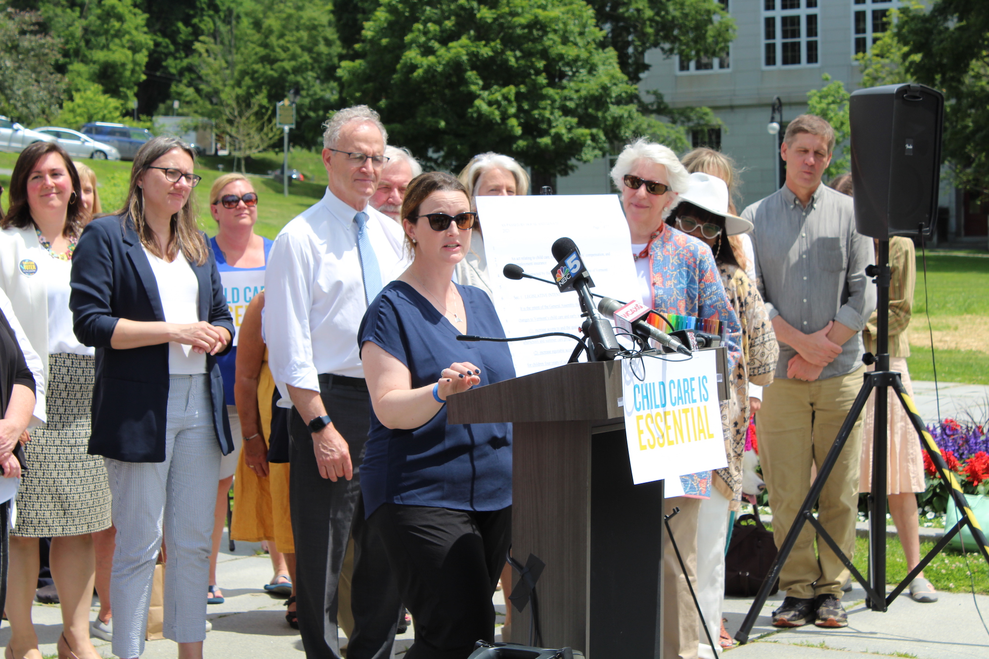 Woman in sunglasses, dark hair, light skin, and a dark blue top stands at a podium speaking outdoors on a sunny day. Behind her, people in business dress listen attentively.