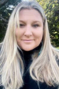 Smiling woman with long blonde hair and light skin with greenery behind her