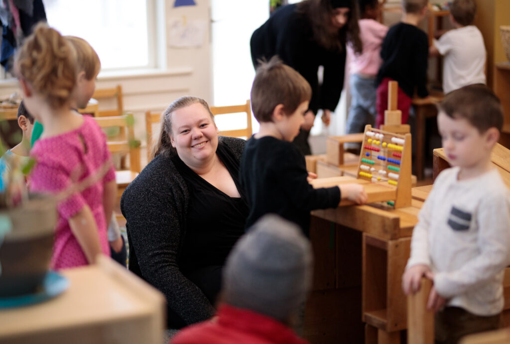 An early childhood educator smiles at the camera in the middle of an active classroom