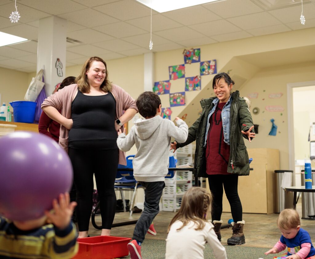 A young boy skips to greet his mother who stands with arms extended while his teacher smiles at them both.