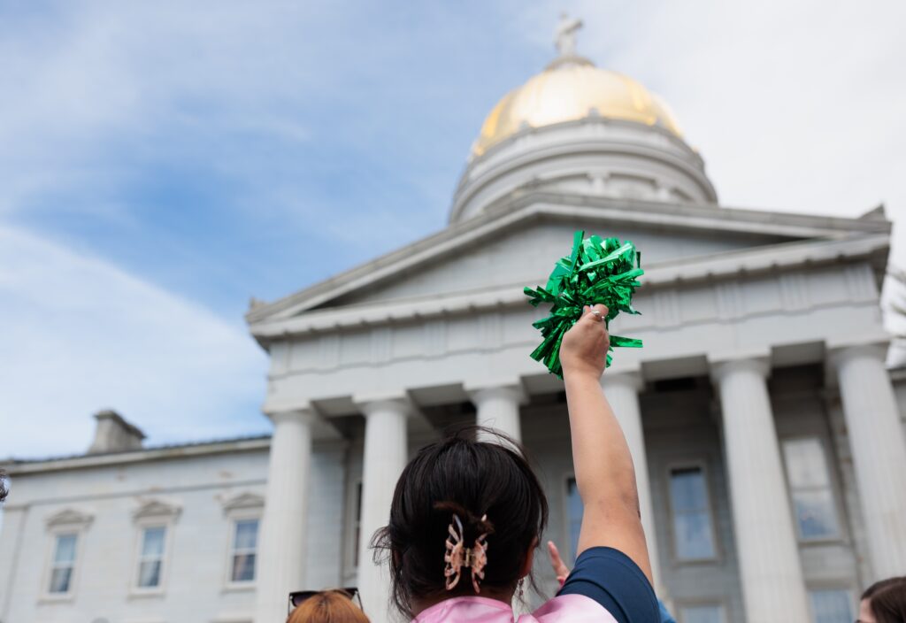 woman with her hair up shakes a green pom pom in celebration in front of the Vermont Statehouse