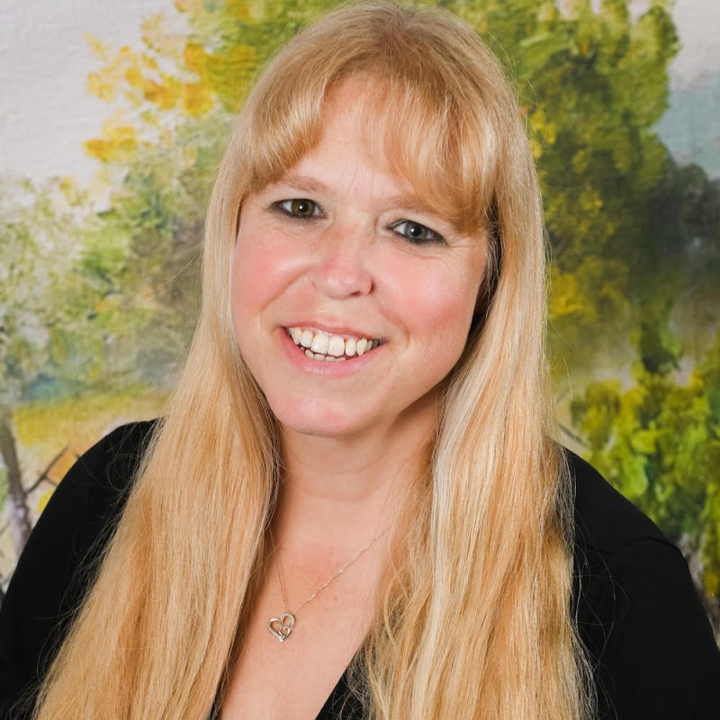 Person with very long blond hair and bangs smiles in front of a painting of trees