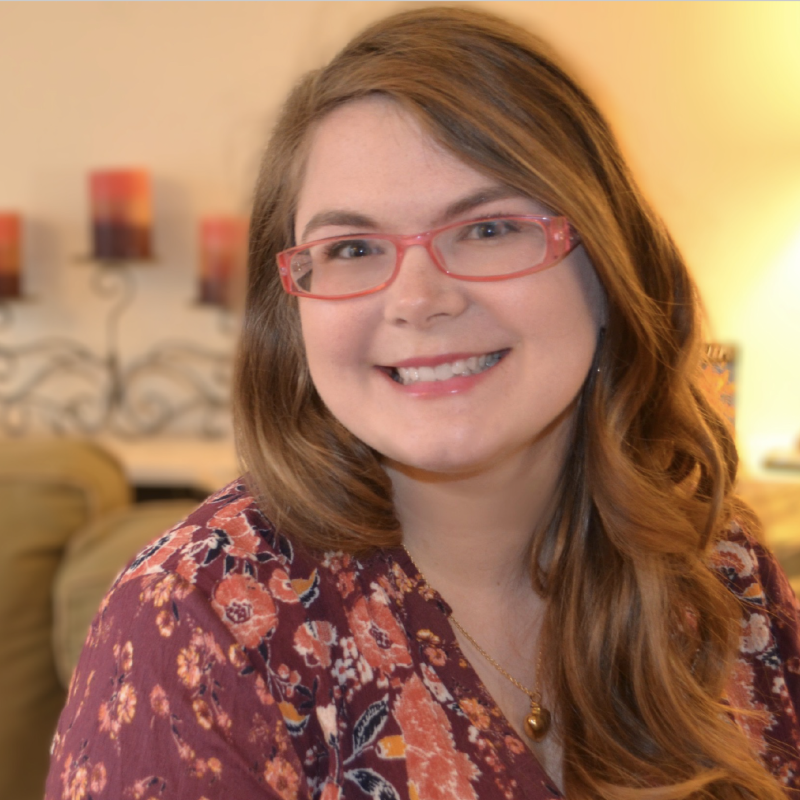 Smiling woman with long wavy brown hair, pink glasses, light skin, floral shirt, indoors